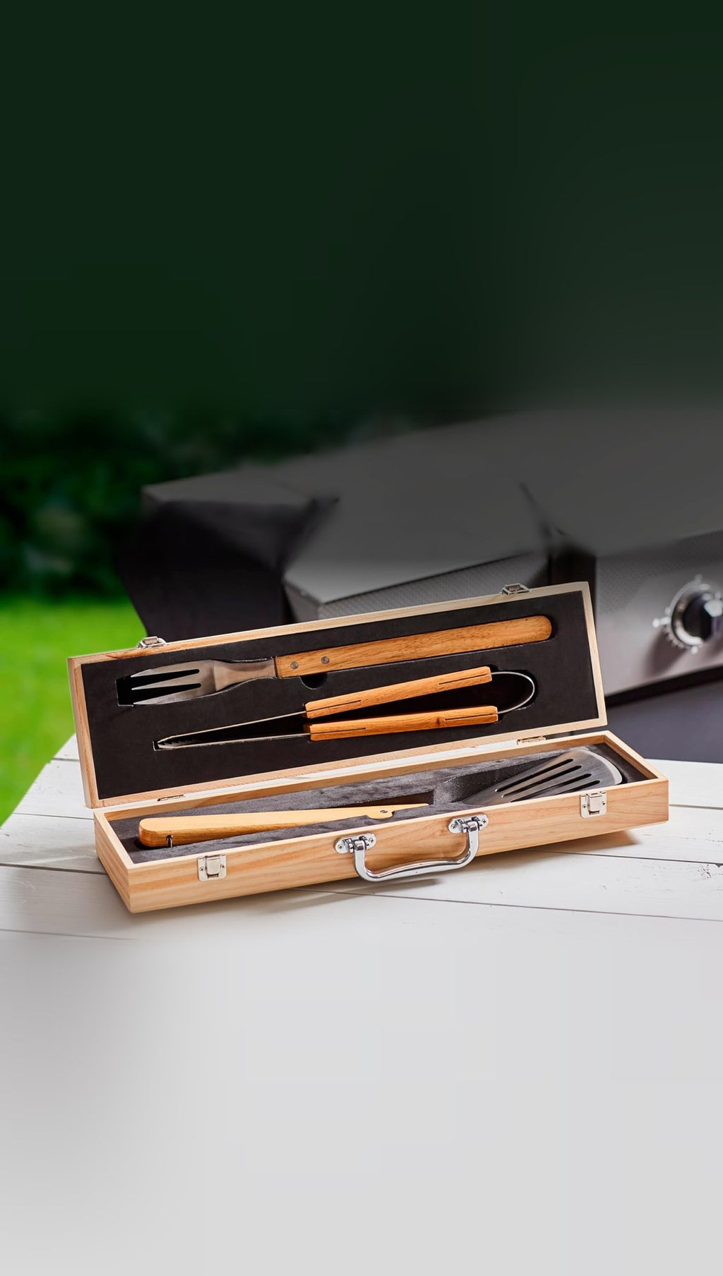 Open pine carry case holding the stainless steel BBQ spatula, fork and tongs with wooden handles, on an outdoor table beside a gas barbecue