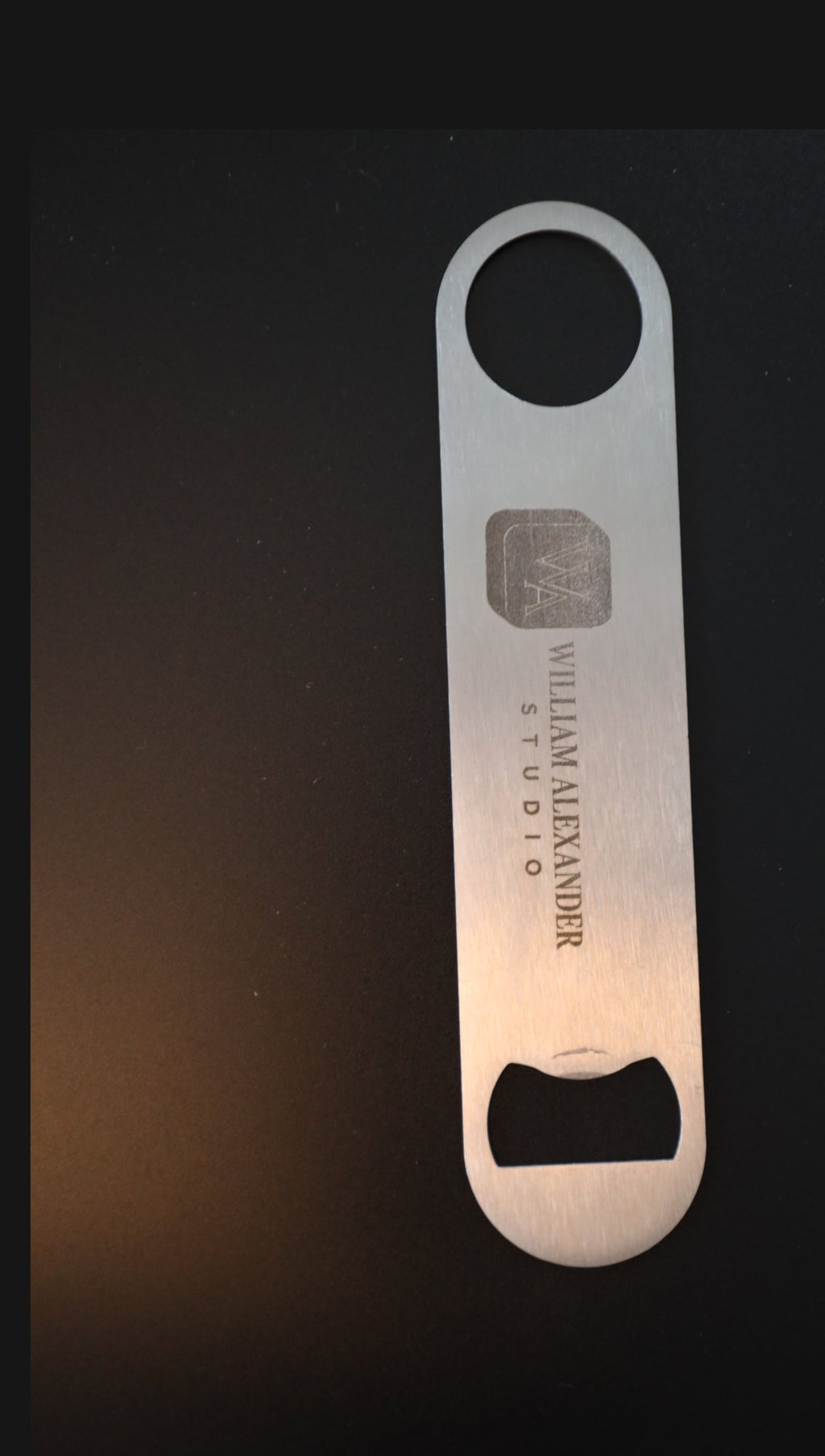 Brushed stainless steel bar blade bottle opener engraved with William Alexander Studio logo, upright on black